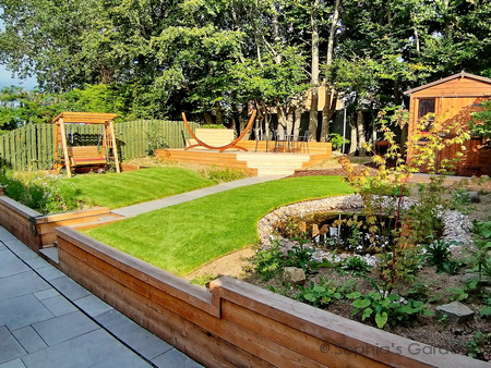 A terraced rear garden with a Siberian larch retaining wall the full width of the garden. Above the wall, to the right, lies a Japanese maple tree, a cobble-bordered wildlife pond and a bespoke wooden shed. To the right are meadow borders, a swing seat and a large deck. Click image for more details.